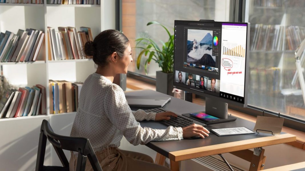Uma mulher está sentada em uma mesa usando um computador Lenovo para trabalhar, encontrando soluções e utilizando vários dispositivos.
