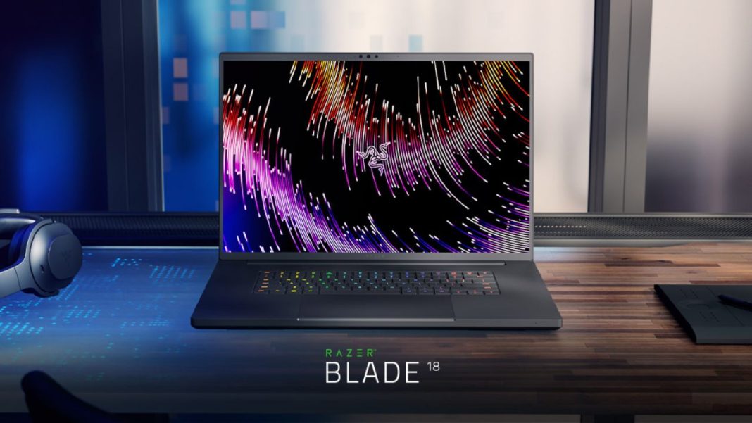 Um portátil gamer Razer Blade 18 mais poderoso com um papel de parede abstrato colorido na tela, colocado em uma mesa com fones de ouvido e um tablet por perto.